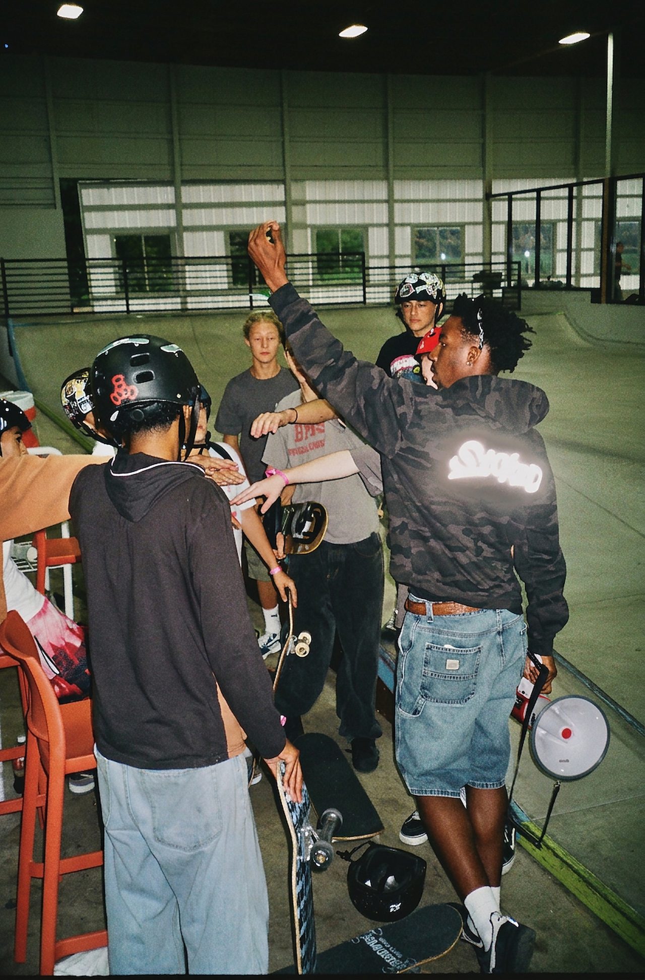 A coach in a STOKED hoodie, one arm raised at the center of a huddle of young skaters in helmets, boards in hand, at an indoor skatepark.