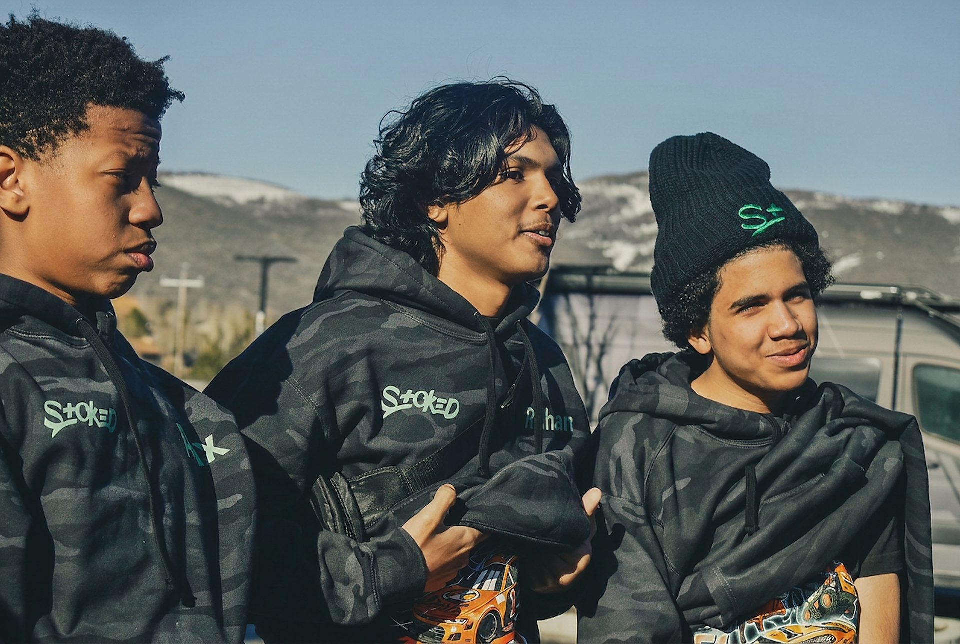 Three young people in black camo STOKED hoodies talking in the sun before a snow session, mountains behind them.